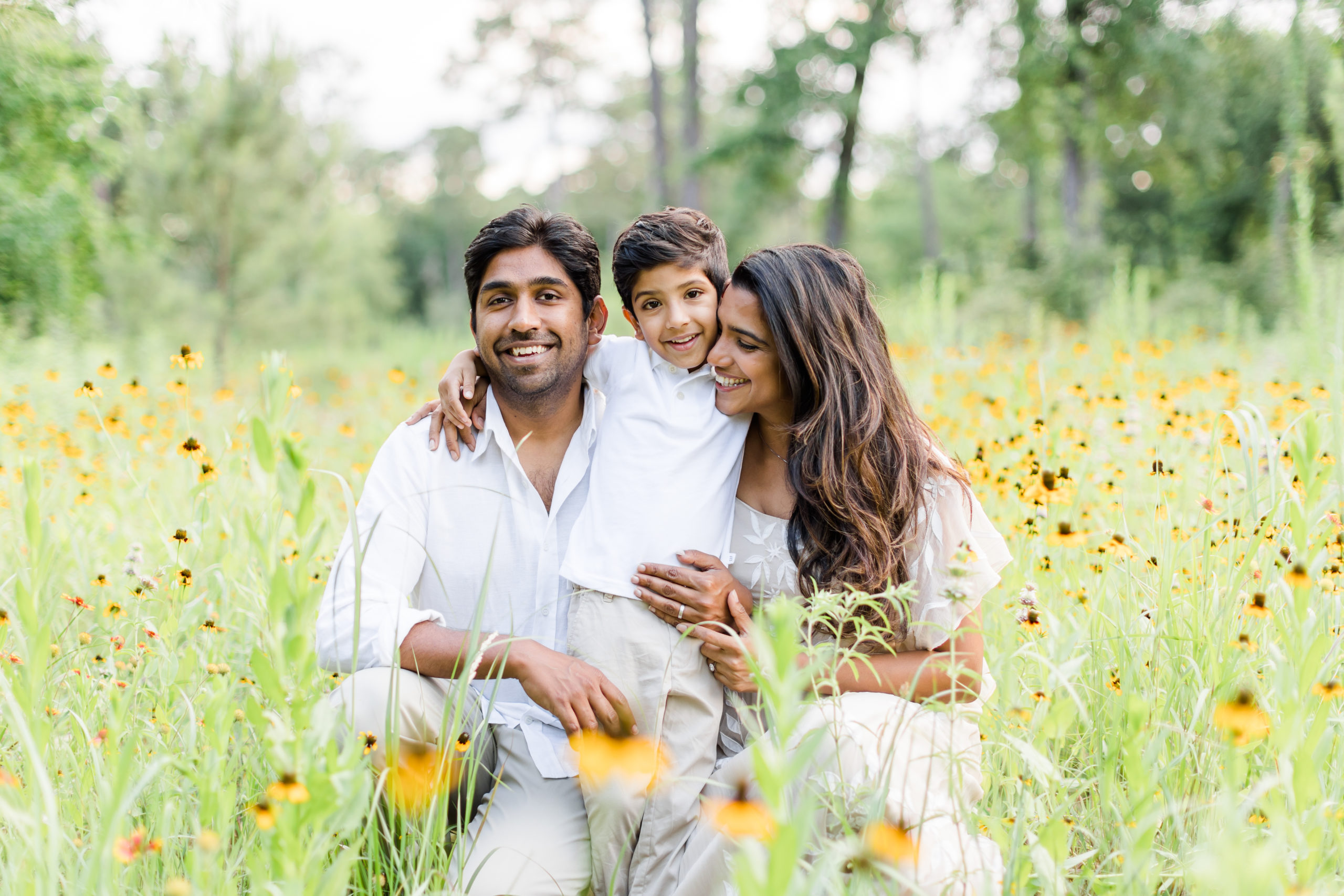Crouched down in a field of yellow wildflowers, Houston, Texas's Monette Anne Photography captures this family of three huddled together and smiling during their family photos. houston texas wildflower sunflower photo session cream and white family photo colors houston texas family photographer #HoustonArboretumandNatureCenter #HoustonTexasFamilyPhotographer #HoustonTexasPhotographer #HoustonHeightsPhotographer #HoustonHeightsFamilyPhotographer #FamilyPhotoOutfitInspiration #NeutralFamilyPhotoOutfits #SummerFamilyPhotos #MonetteAnnePhotography #FamilyPhotoPoses