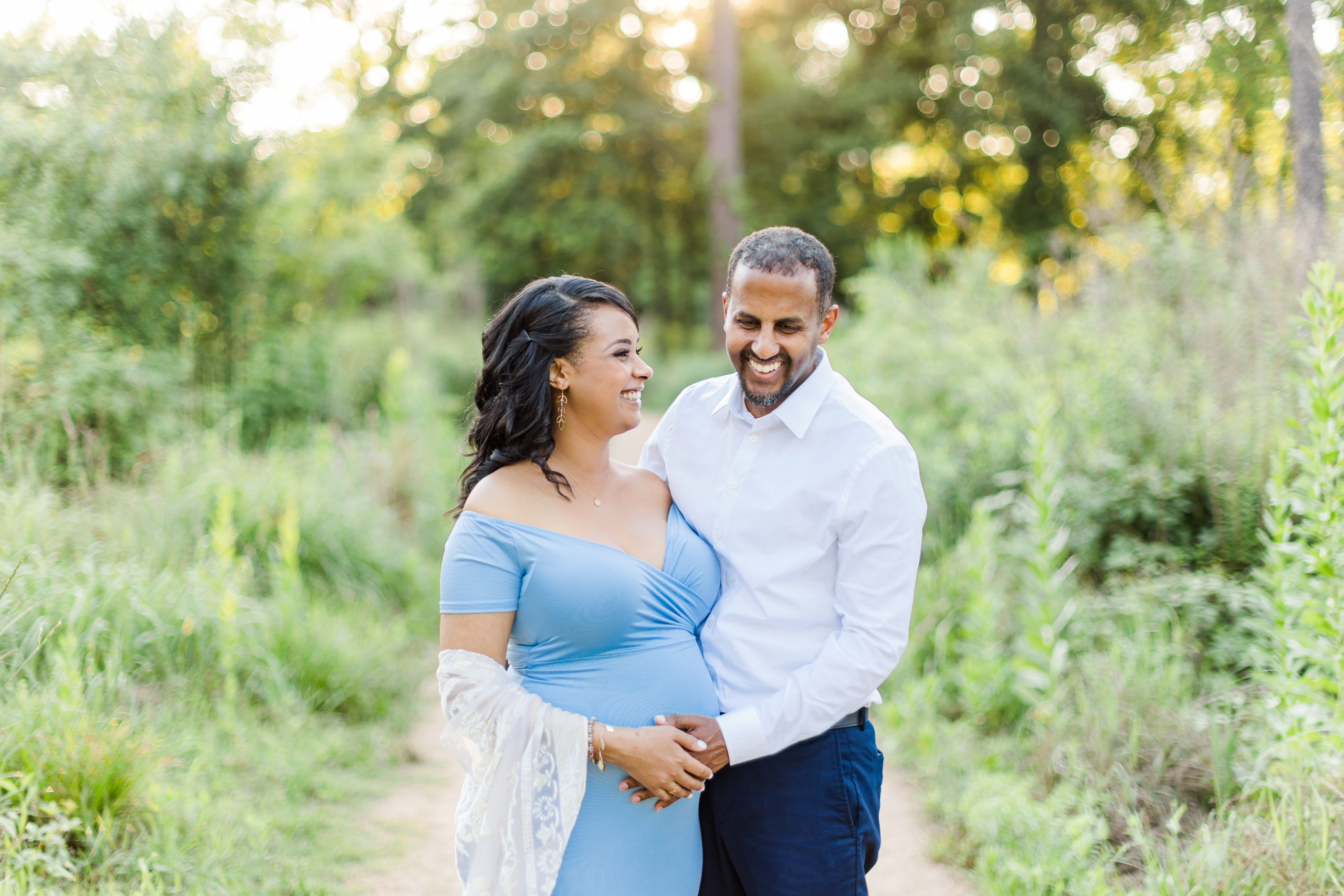 During this maternity session in Houston Heights, Texas, Monette Anne Photography captures this expectant couple posing happily together. expectant father holding wife's stomach baby bump fitted blue off the shoulder womens maternity dress white lace womens shawl gold womens jewelry #HoustonArboretumandNatureCenter #HoustonTexasMaternityPhotographer #HoustonTexasCouplesPhotographer #HoustonHeightsMaternityPhotographer #HoustonHeightsCouplesPhotographer #TexasGalleriaPhotographer #MaternityPhotos #BrightAndAiryPhotography #MaternityDress #SummerMaternityPhotos