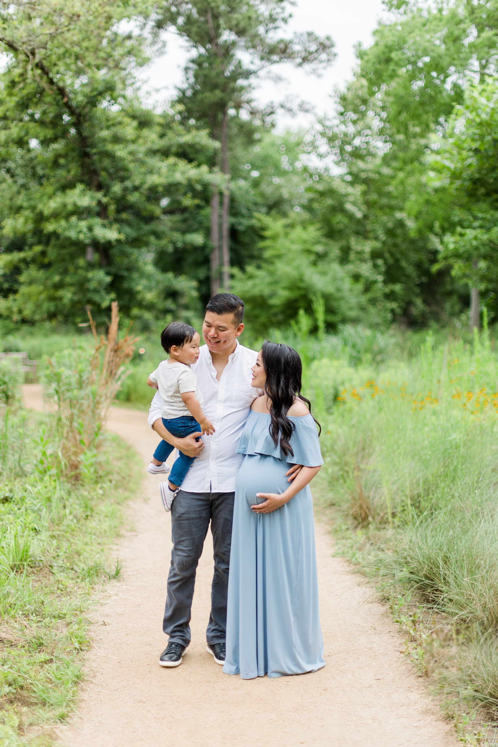 On an unpaved road in Houston, Texas, this family of three pose together happily for their maternity session with Monette Anne Photography. family maternity session womens off the shoulder maxi maternity dress pregnancy announcement twin mom baby bump houston texas photographer #HoustonArboretumAndNatureCenter #HoustonTexasMaternityPhotographer #HoustonTexasFamilyPhotographer Houston #HoustonHeightsPhotographer #MonetteAnnePhotography #HoustonTexasPhotographer #MaternityPhotoShoot #PregnancyAnnouncement #BabyBump #ExpectantParents