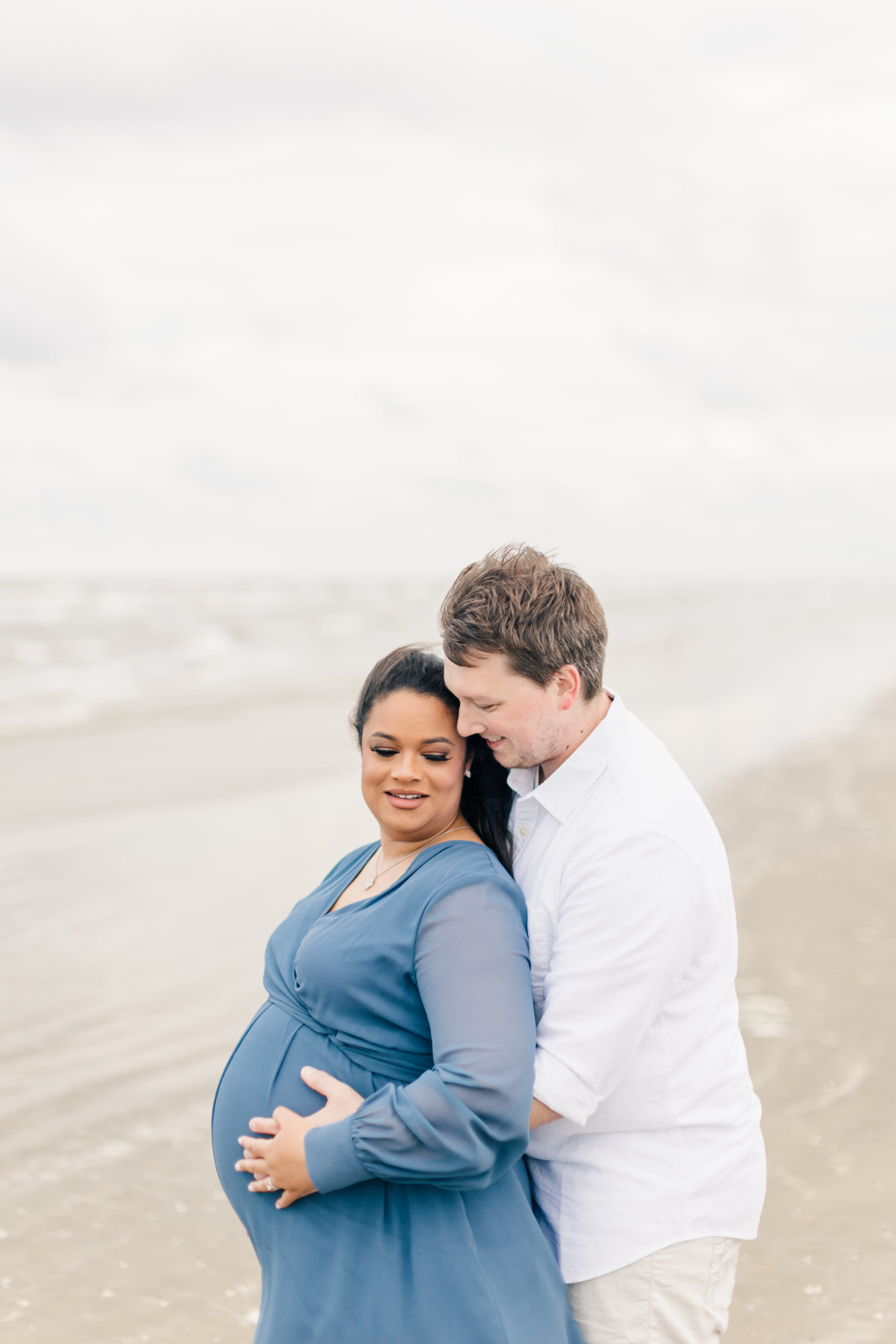 During this beach maternity session held at Galveston Beach, Monette Anne Photography captures this soon-to-be father hold his pregnant wife from behind. man hugging wife from behind gray blue long sleeve maternity dress beach maternity session houston texas #GalvestonBeach #HoustonTexasMaternityPhotographer #HoustonTexasCouplesPhotographer #PiratesBeachTexas #MaternitySession #MonetteAnnePhotography #GalvestonBeachMaternitySession #MaternityClothes #BeachMaternitySession