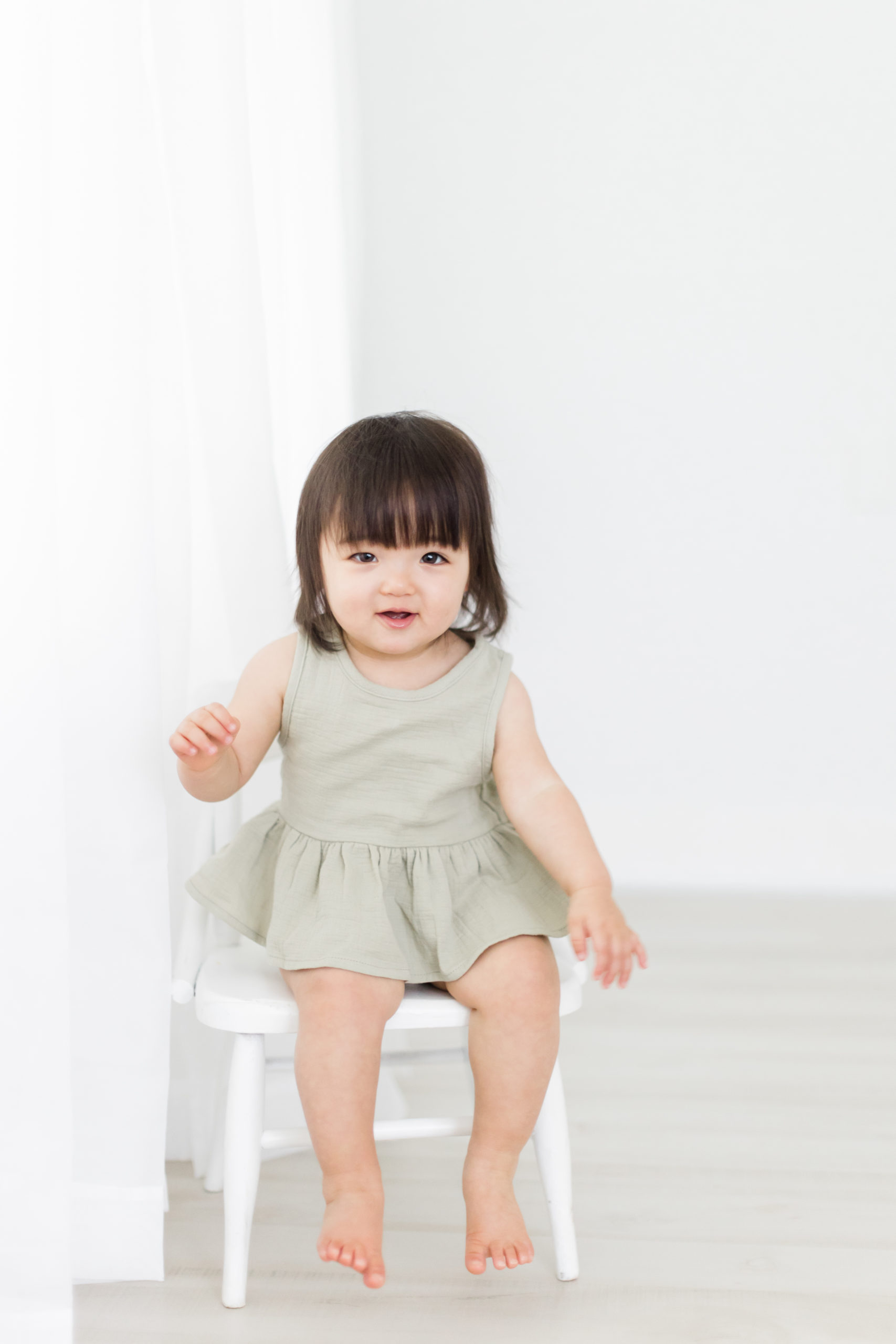 At a natural light photography studio in Houston, Texas, Monette Anne Photography captures this toddler girl sitting on a white wooden stool. toddler sitting on stool olive green toddler dress houston photographer #HoustonTexasFamilyPhotographer #MontroseTexasFamilyPhotographer #MuseumDistrictTexasPhotographer #WestUniversityTexasPhotographer #HoustonPhotographyStudio #MonetteAnnePhotography #HoustonTexasPhotographer #FirstBirthday #MontroseTexasPhotographer #SmashCake