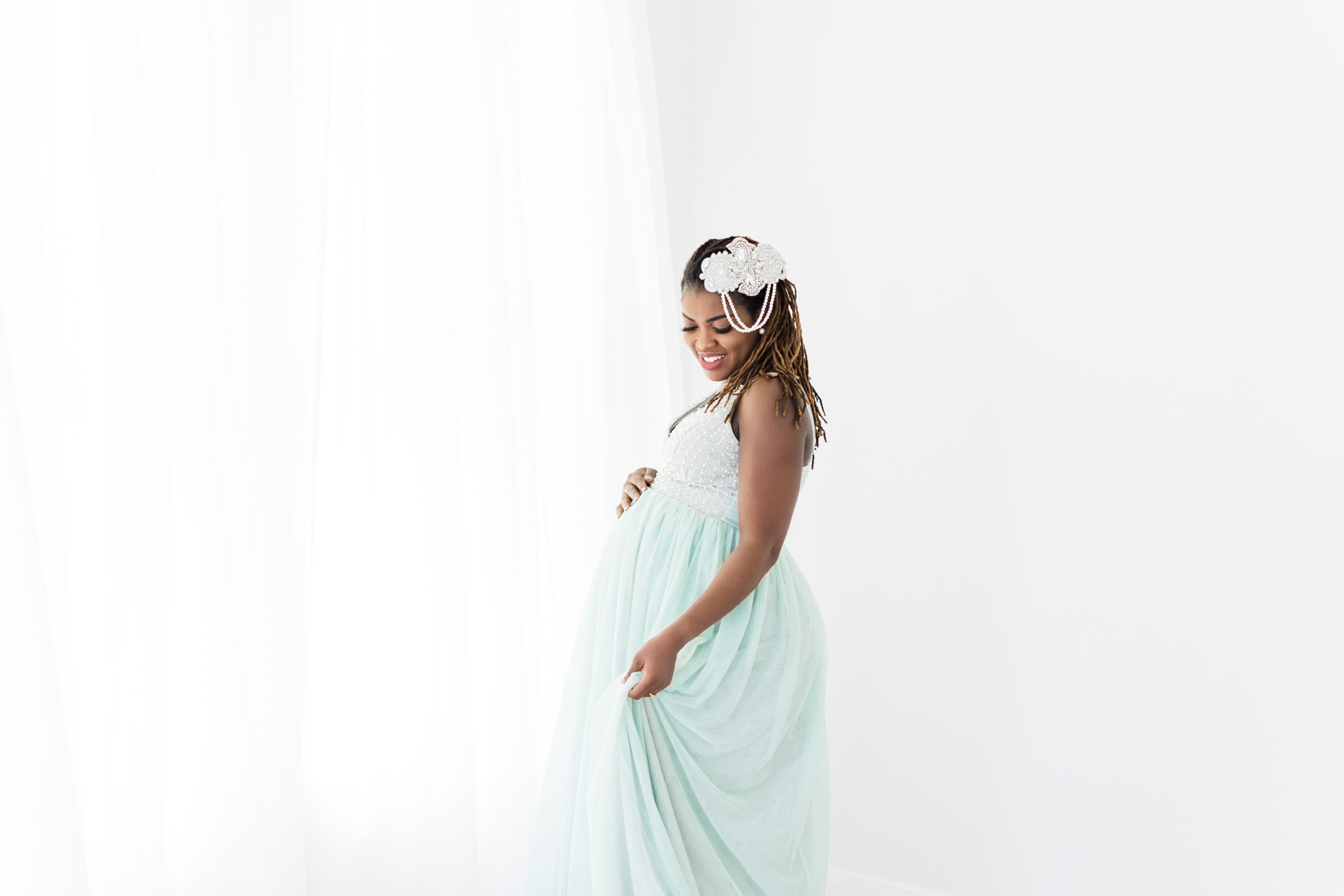 Monette Anne Photography captures a stunning black mother with light blue gown and pearl hair clip smiling while looking down in the Houston area. soon to be mother texas houston maternity photographer #monetteannephotography #monetteannematernity #houstonmaternityphotographer #houstonphotographers #brightlightstudiophotography #houstonmaternityphotography #maternitystyle #babybump #bumppictures #parentstobe #maternitydress #whitestudio #texashoustonphotographers #babyontheway
